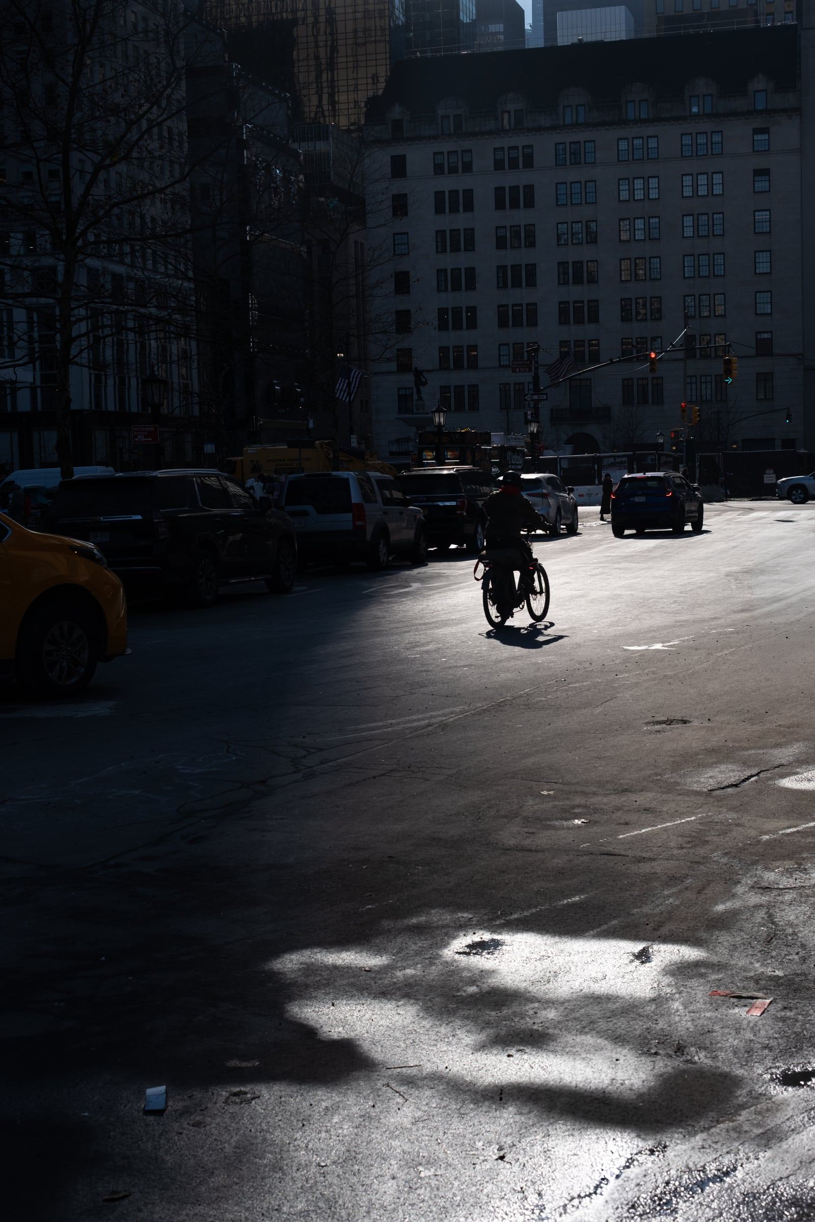 A cyclist rides through a single shaft of sunlight on a shadowed Manhattan street.