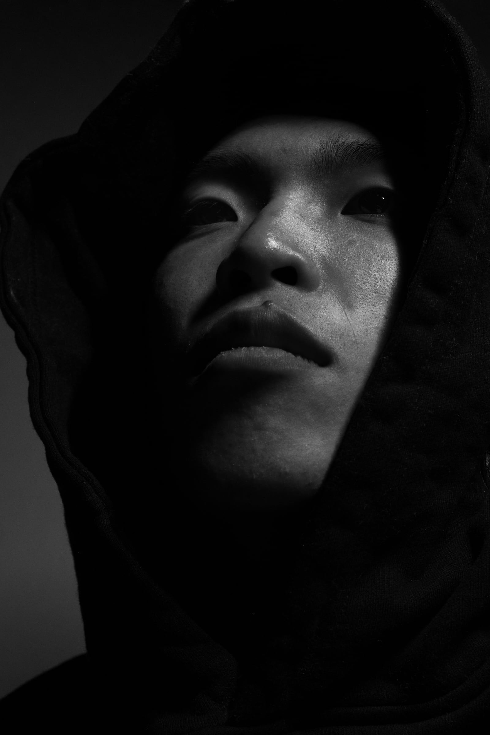 Black-and-white portrait of a young man in a hood, his face half-lit against darkness.