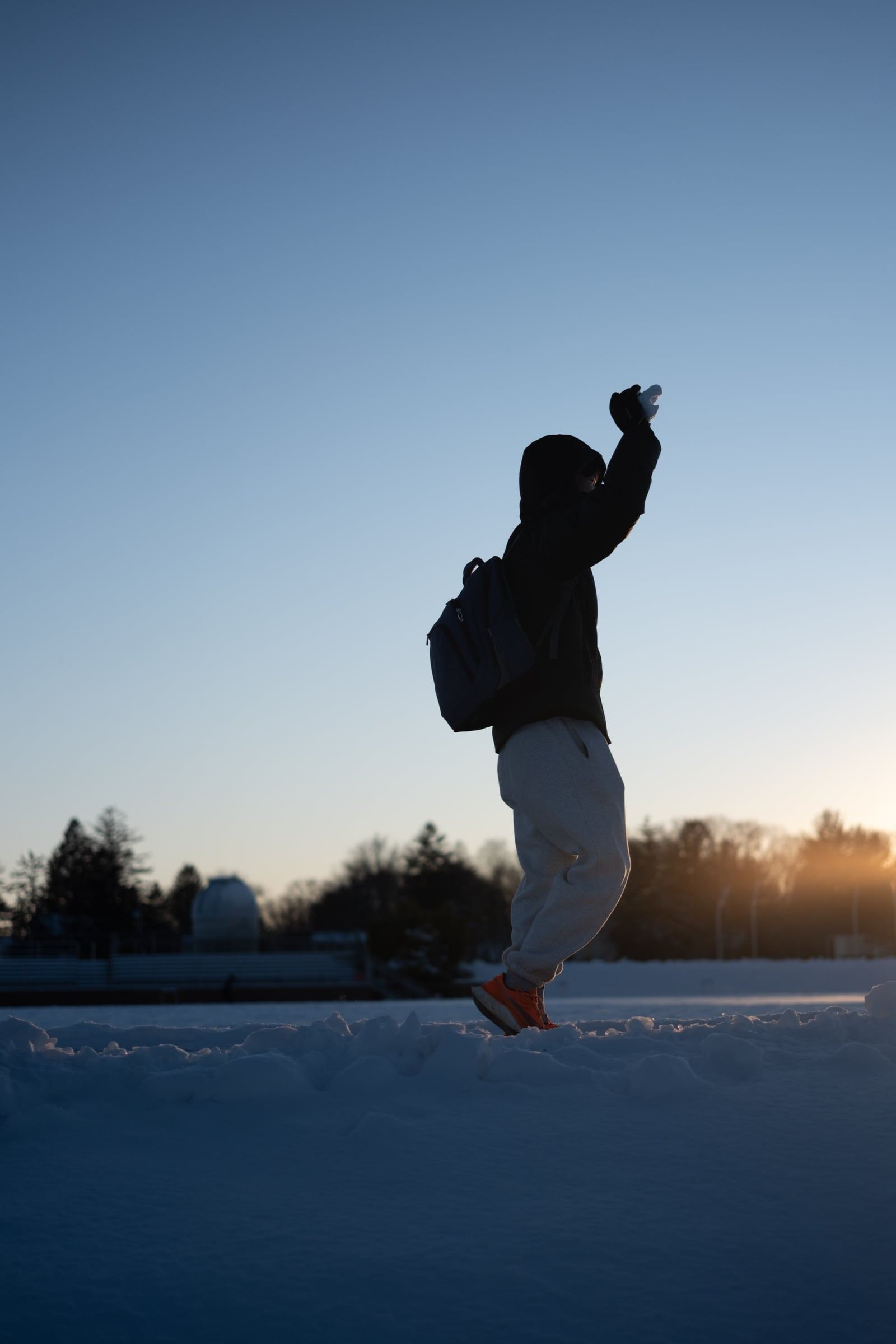 A silhouetted figure with a backpack leaps on a snowfield, arm raised against the evening sky.