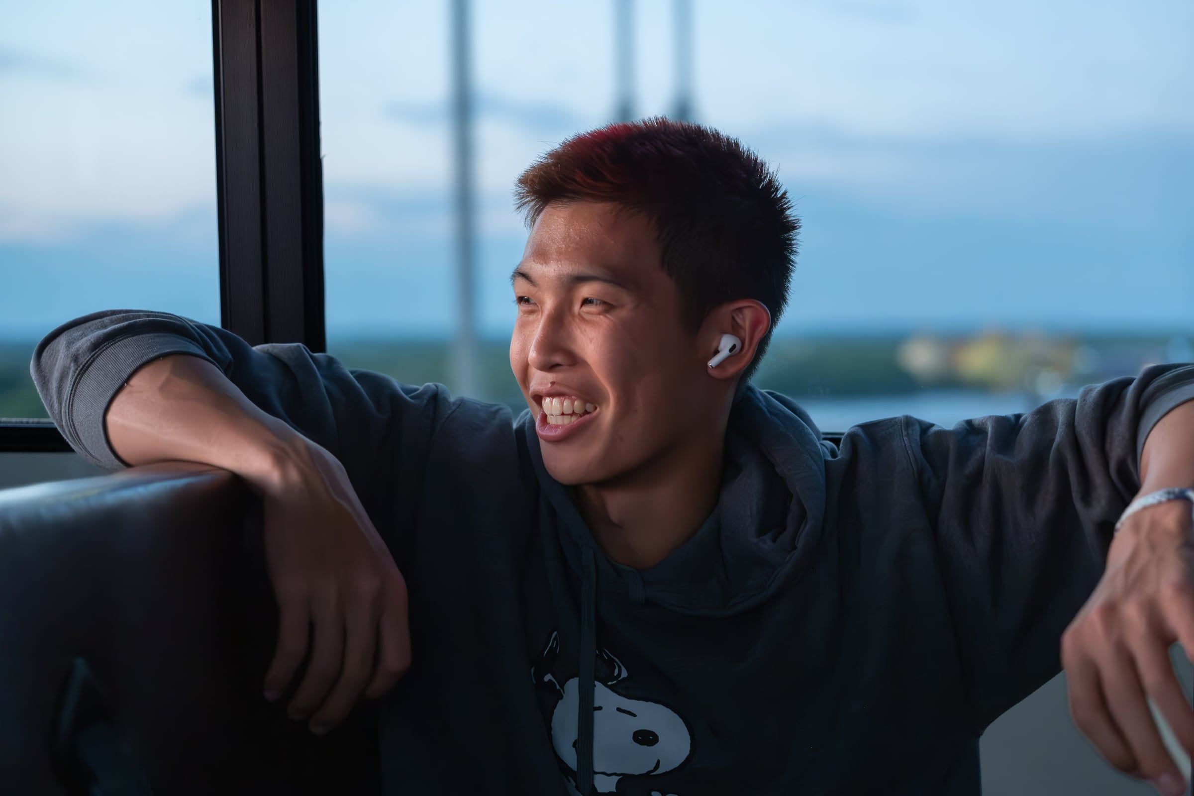 A young man in a hoodie laughs, leaning on a sofa by a window at dusk.
