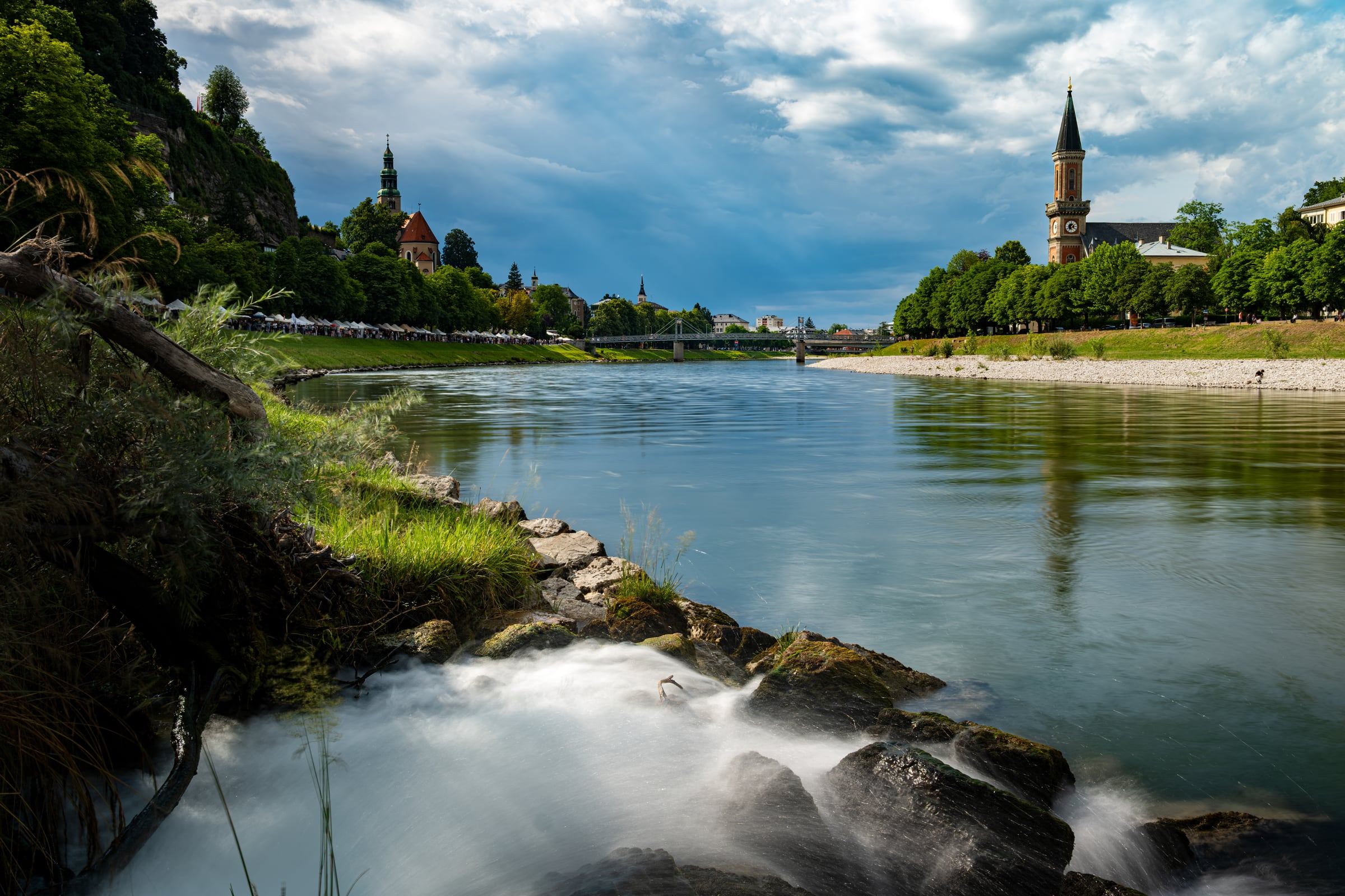 Silky water rushes over rocks in the foreground of a wide river lined with trees and church spires.