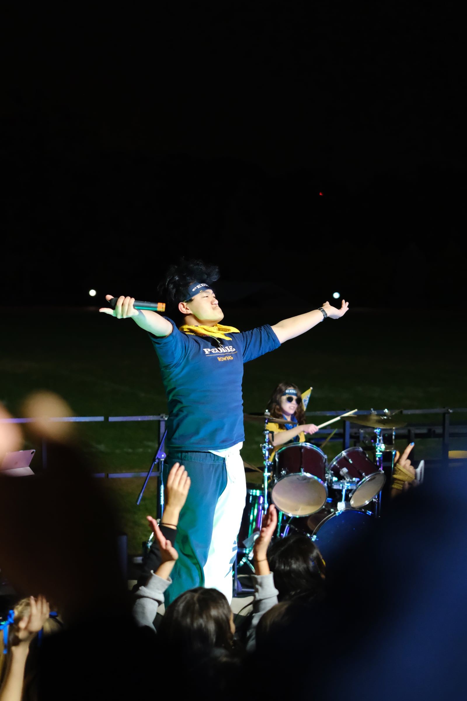 A singer stands on an outdoor stage at night, arms spread wide, as the crowd raises hands.