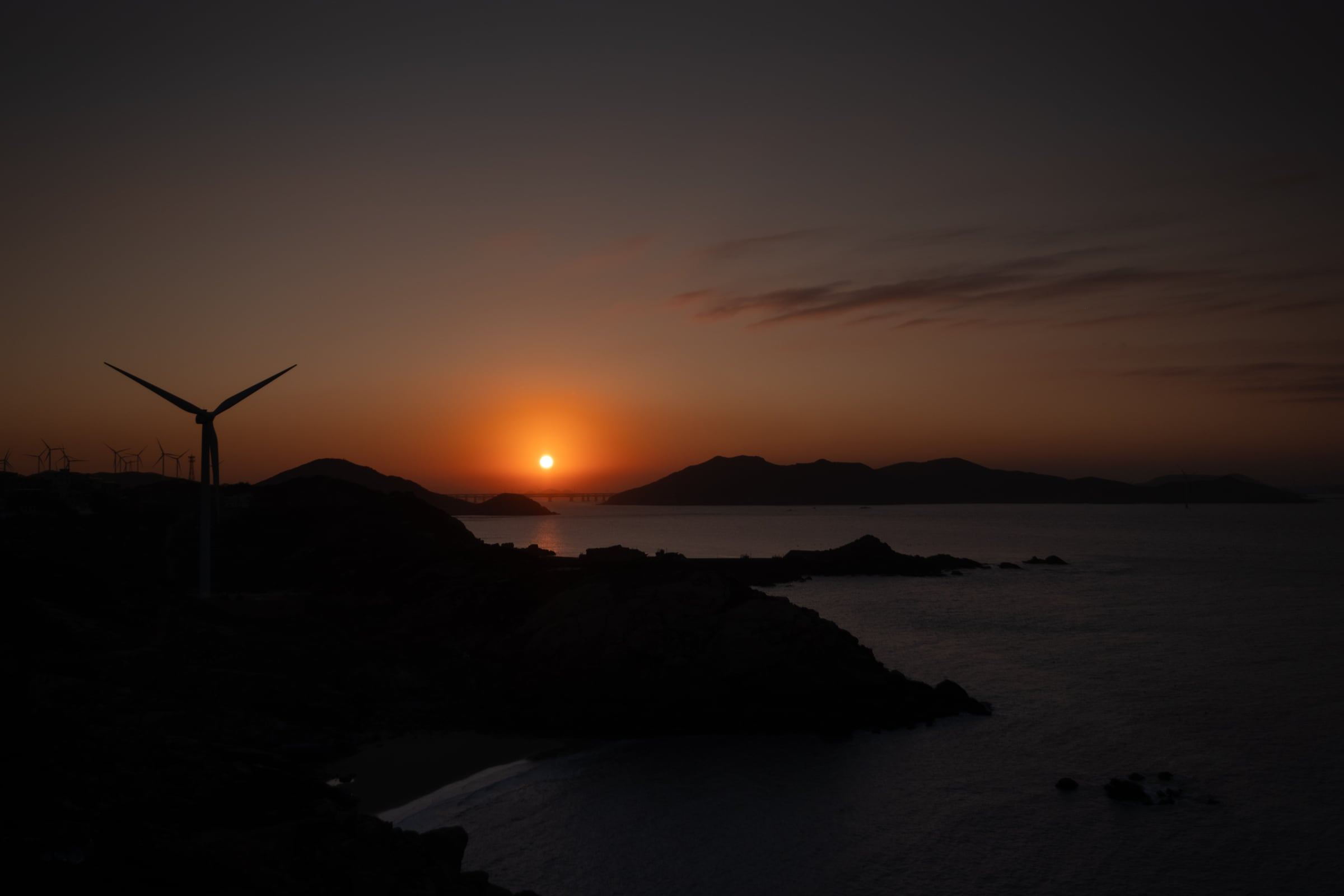 The sun setting between dark island silhouettes, with a lone wind turbine on the headland.