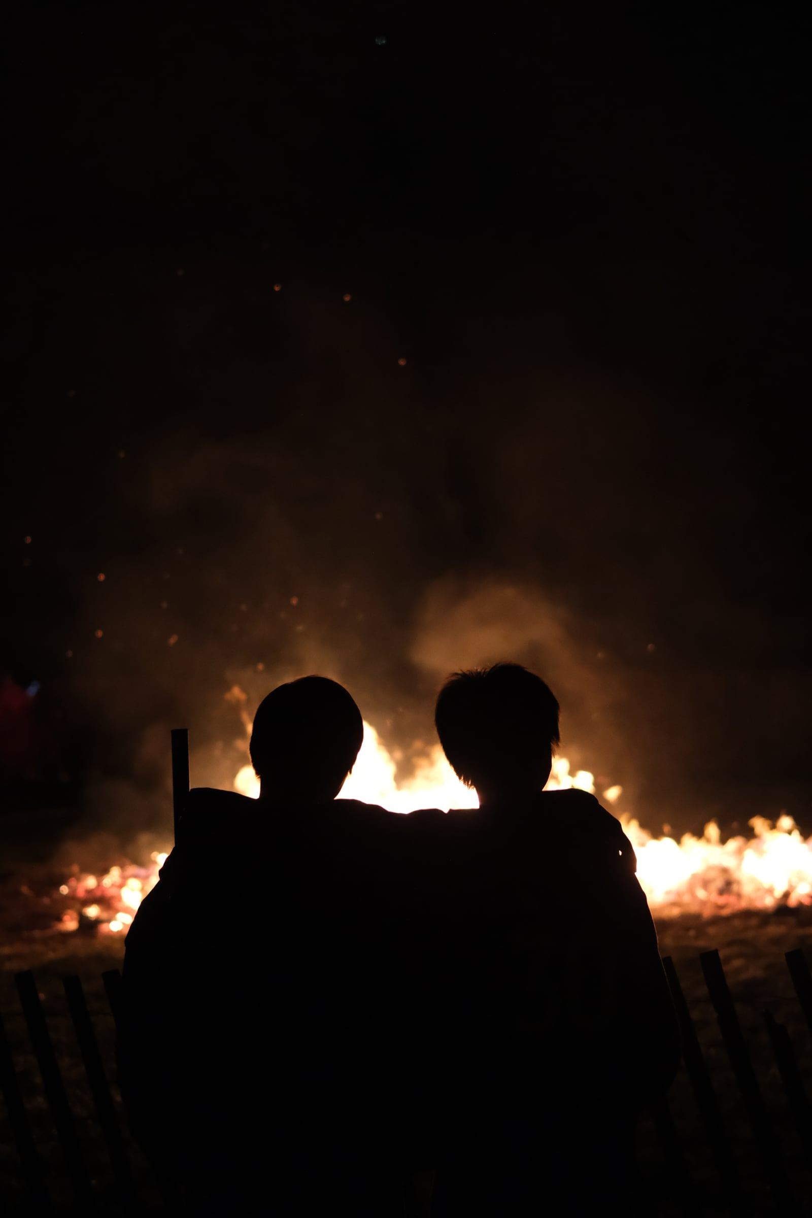Two silhouetted figures sit side by side in front of a large bonfire at night.