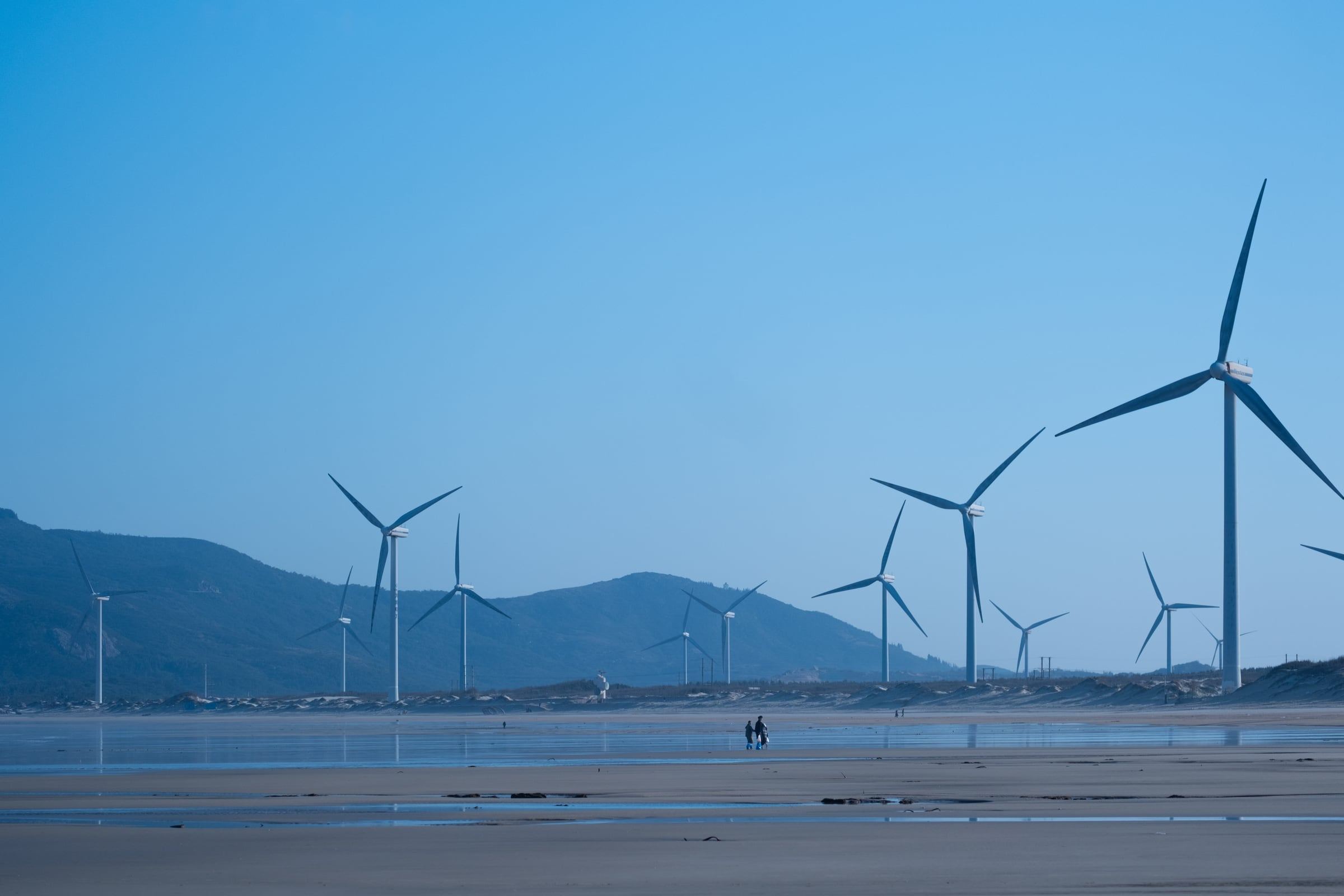 Two small figures walk across a wet beach below a field of wind turbines and blue hills.